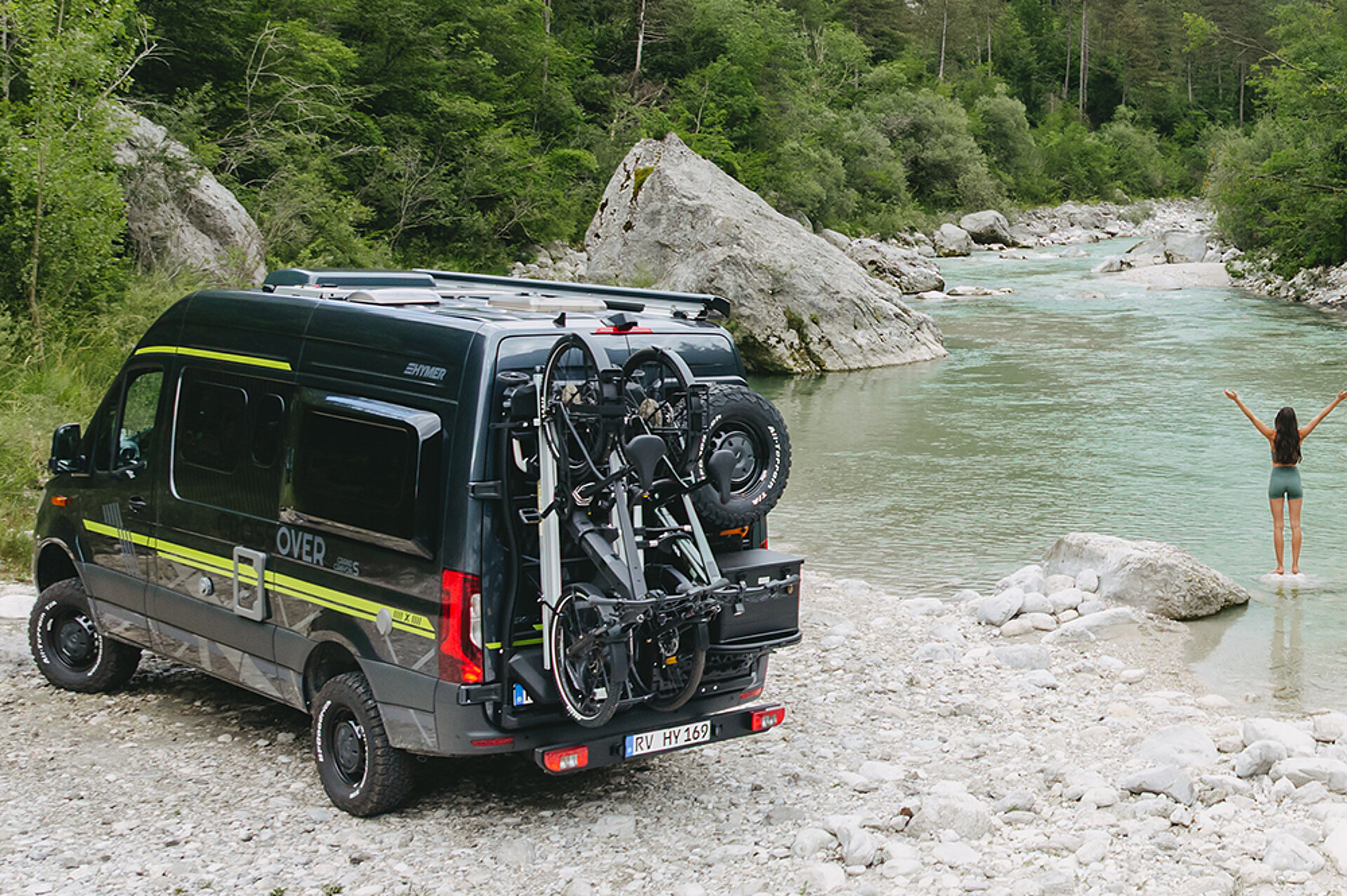 Abenteuer inklusive – mit den Sondereditionen des Grand Canyon S. Ein HYMER Grand Canyon S CrossOver steht an einem Bergsee, eine Frau steht davor und breitet ihre Arme aus.