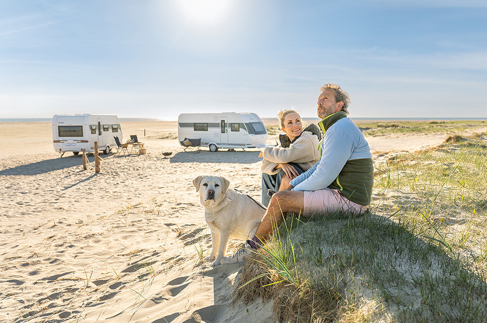 Ein Paar mit Hund sitzt am Strand, im HIntergrund stehen zwei Hobby ONTOUR Wohnwagen.