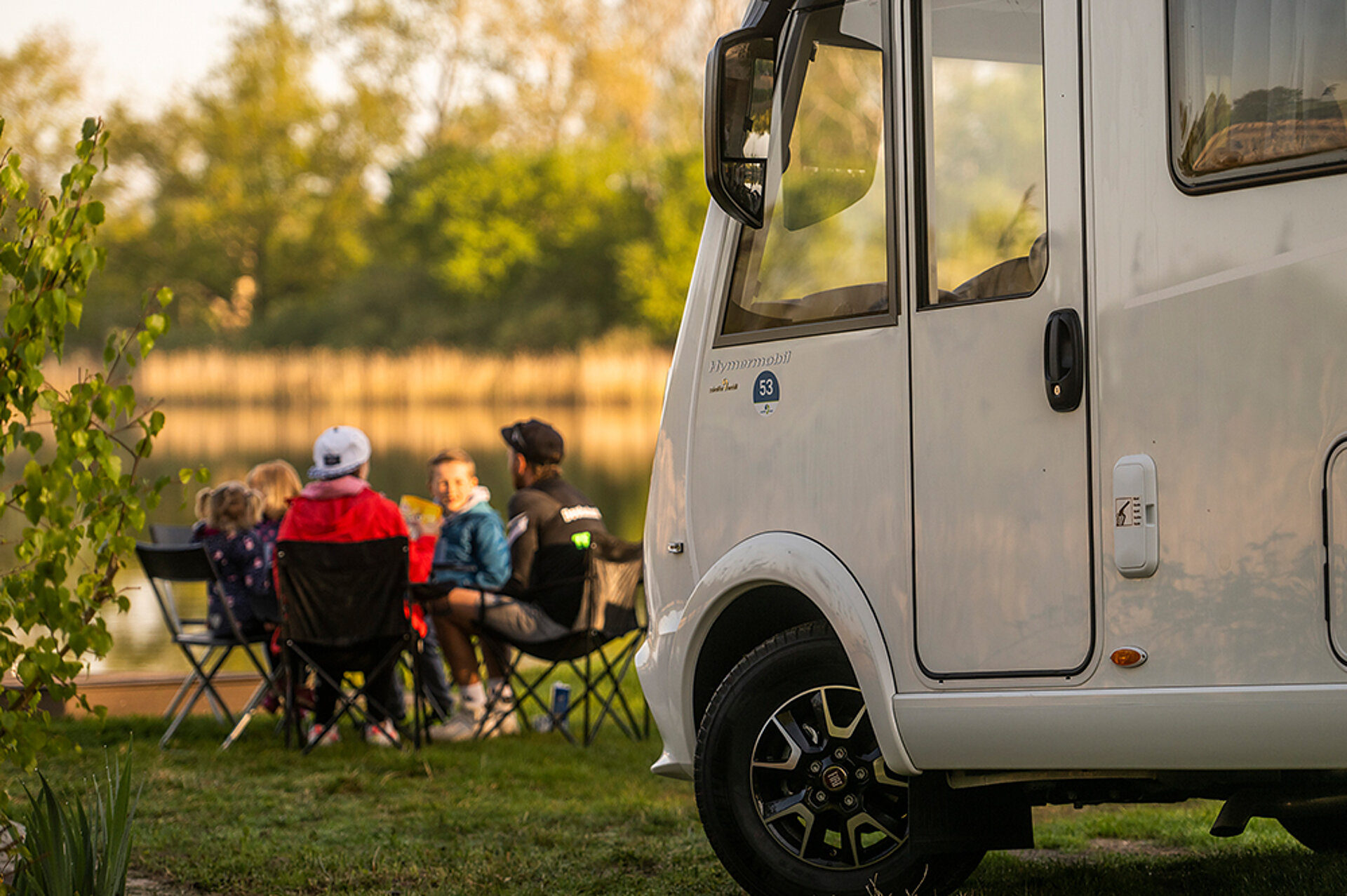 U jezera stojí obytný vůz, rodina sedí na kempingových židlích.