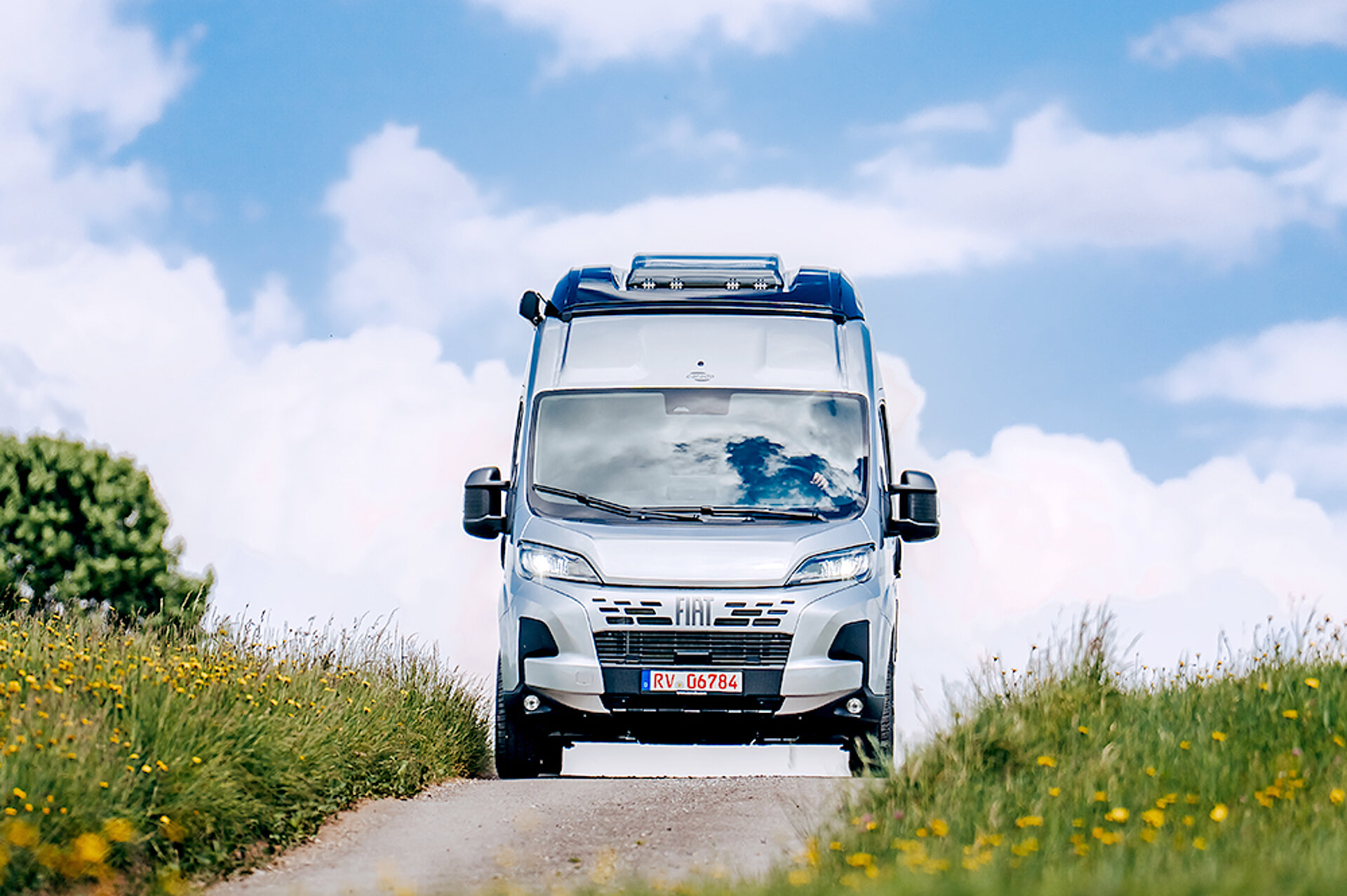 Ein Carado Camper Van fährt über eine Landstraße, die durch eine Wiese führt.
