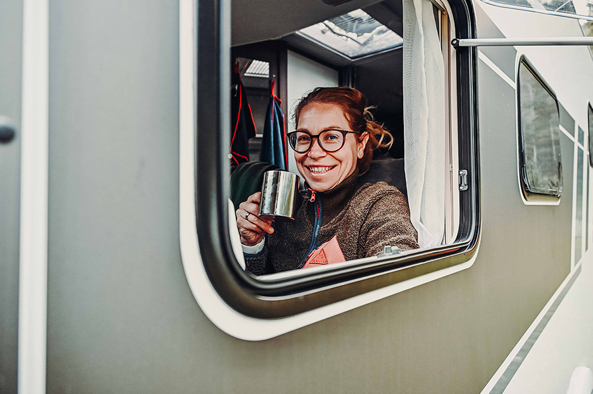 Žena pije kávu a dívá se z okna obytného vozu.