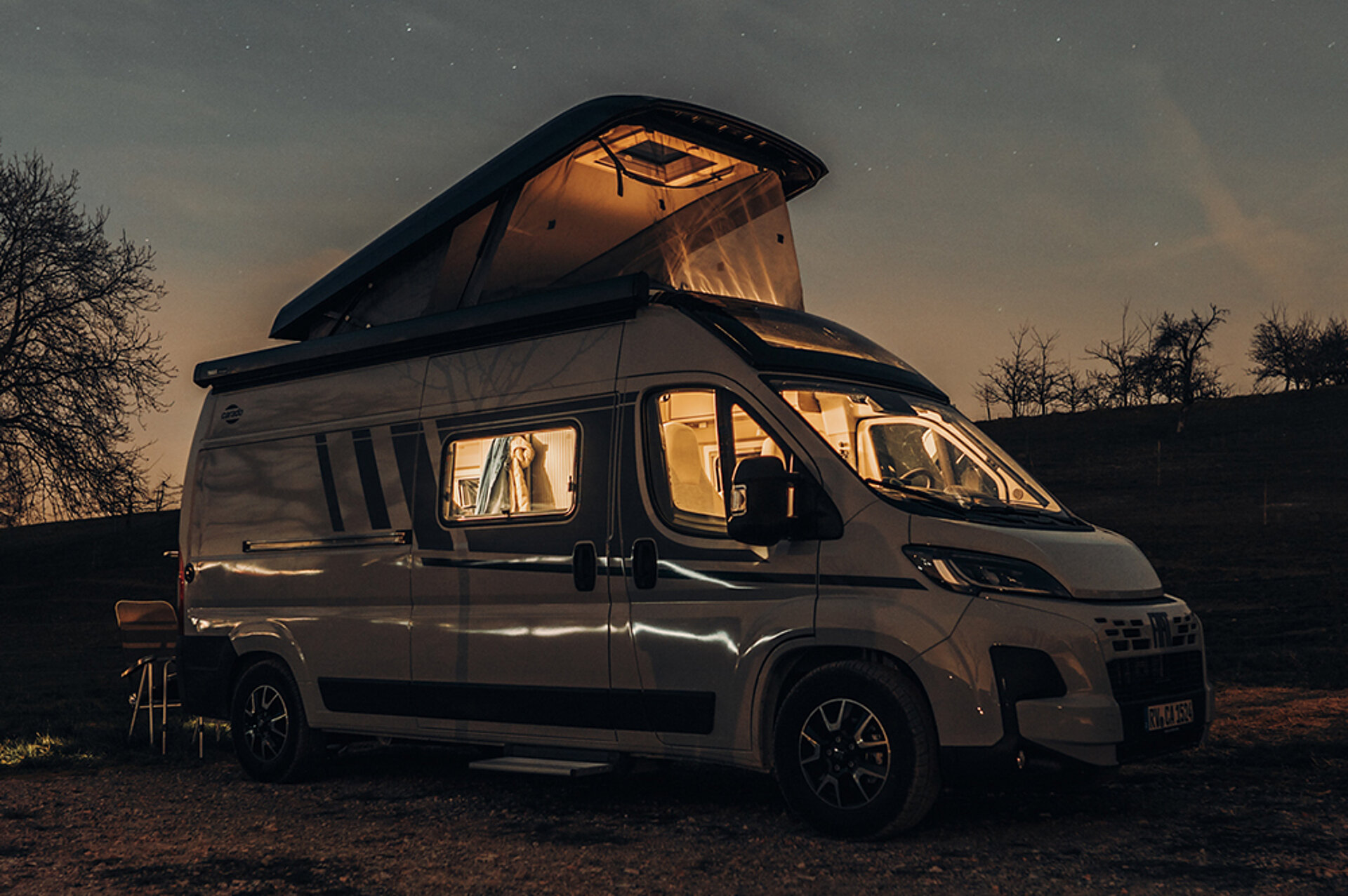 Osvětlený obytný vůz Carado se zvedacím střechou stojí v noci na parkovišti.