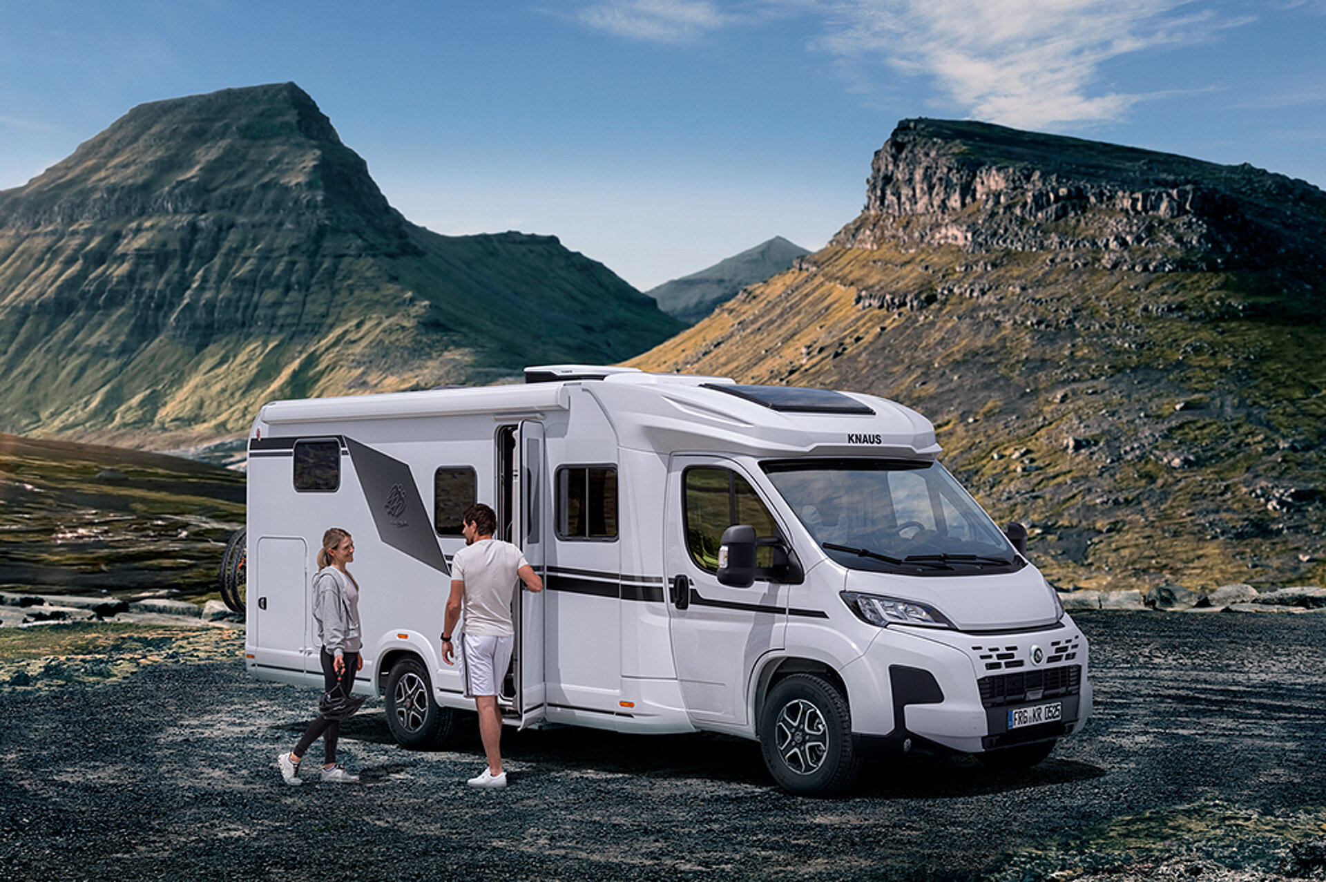 A couple are standing in front of their KNAUS motorhome amidst a picturesque mountain landscape.