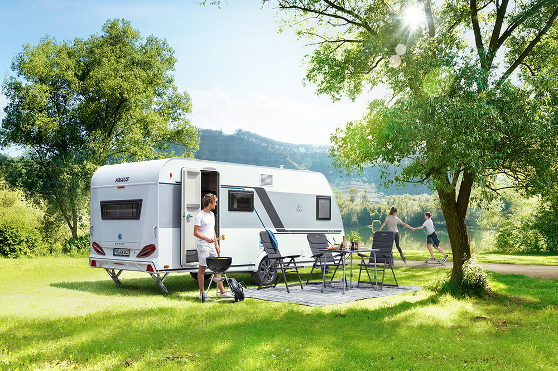 Eine Familie spielt und grillt vor einem im Grünen geparkten Wohnwagen.