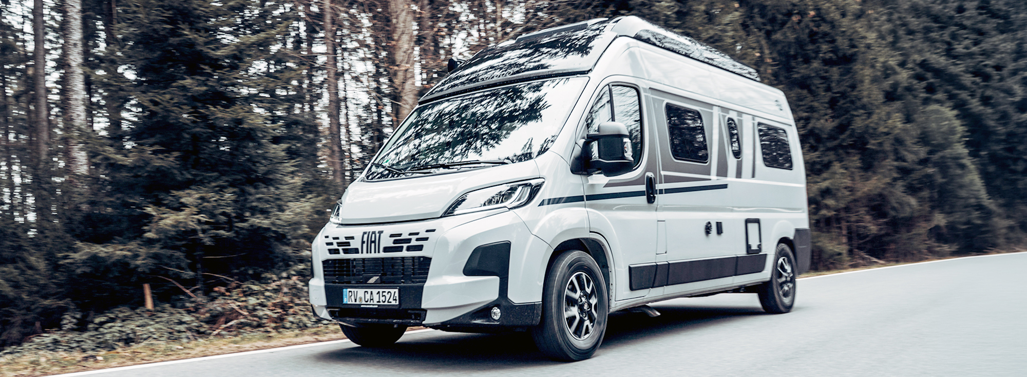 Dodávka značky Carado jede po silnici, která vede lesem.