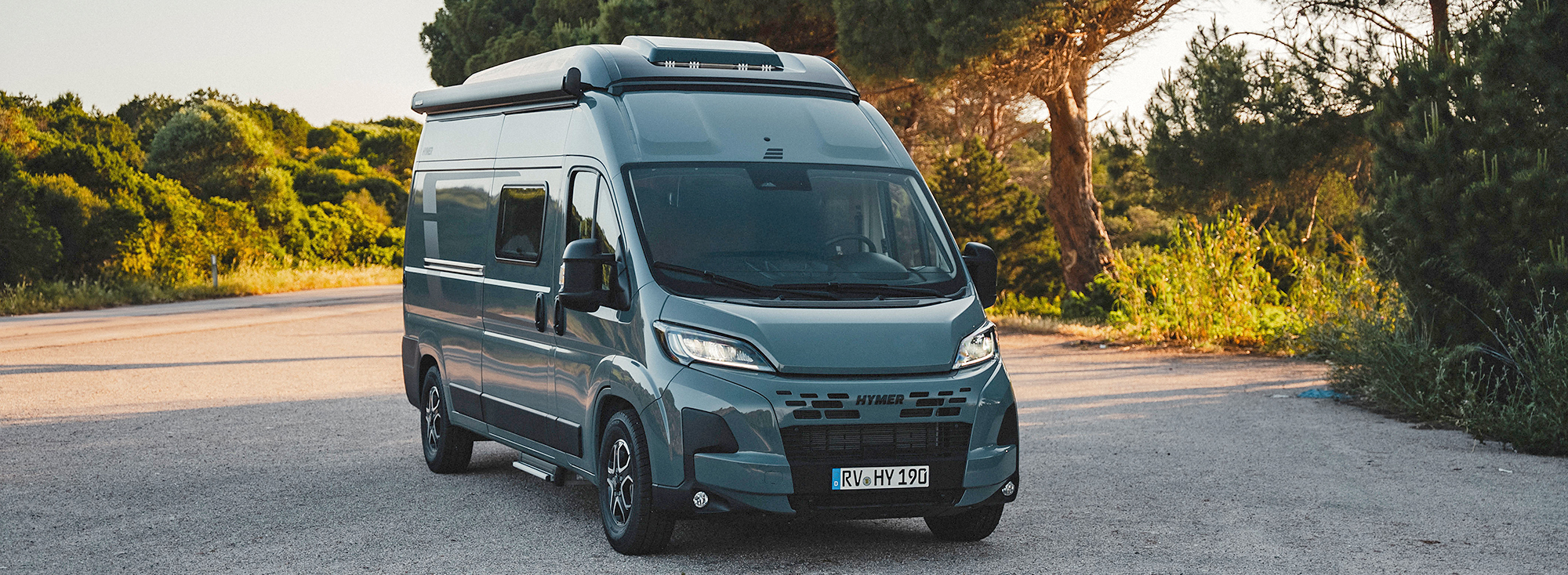 Ein Camper Van steht vor einem Wald im Schatten.