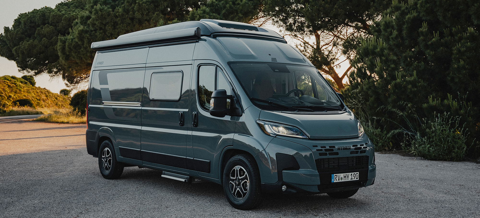 Ein HYMER Redwood Camper Van steht auf einem Parkplatz vor einem Wald. 