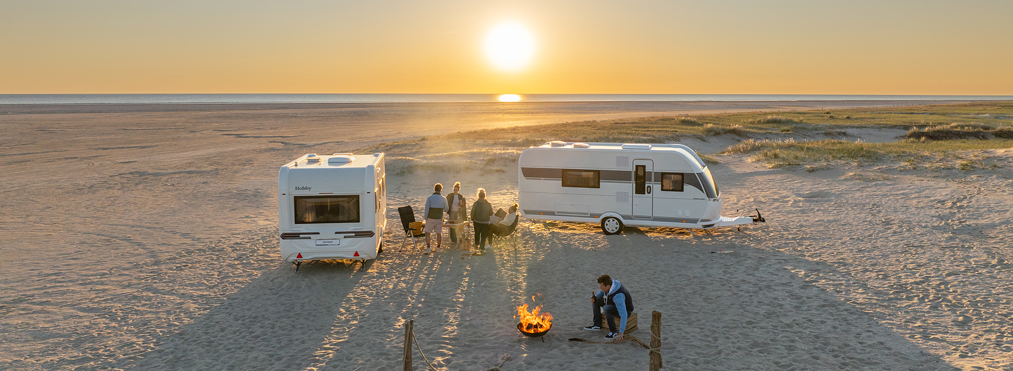Zwei Hobby ONTOUR Wohnwagen stehen am Strand, Freunde grillen davor.
